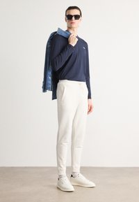 Camisa de manga larga azul marino, pantalones de chándal blancos y zapatillas blancas. Chaqueta de mezclilla colgada del hombro. Gafas de sol que aportan un toque moderno.