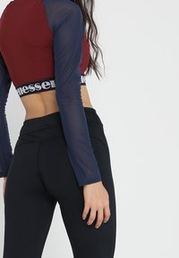 Burgundy crop top dengan lengan sheer biru tua yang panjang dan legging hitam yang memiliki pinggang berlogo, tekstur halus, dan desain yang pas.