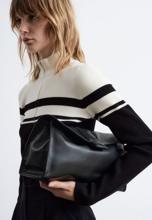 Schwarze Ledertasche mit strukturiertem Design, ausgestattet mit einem flachen Boden und einem umschlagbaren Deckel. Getragen von einem Modell mit einem gerippten, gestreiften Pullover.