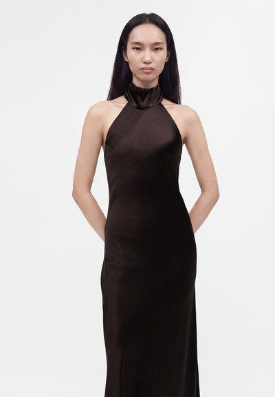 Femme aux longs cheveux noirs porte une robe noire sans manches à col haut, debout les mains derrière le dos sur un fond blanc uni.