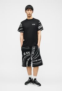 Νεαρός άντρας στέκεται με τα χέρια στις τσέπες, φορώντας μαύρο υπερμεγέθες μπλουζάκι και μαύρο σορτς με μεγάλα λευκά σχέδια κειμένου, λευκές κάλτσες και μαύρα αθλητικά παπούτσια.