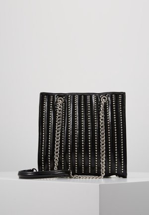 Czarna skórzana torebka z pionowymi srebrnymi ćwiekami i srebrnymi łańcuchowymi paskami na ramię, na białej powierzchni na tle szarego tła.