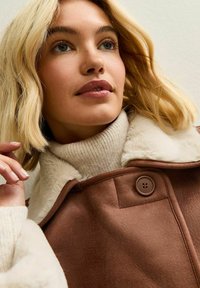Cappotto con collo in shearling marrone e chiusura a bottone, abbinato a un maglione a coste crema. Il modello ha capelli biondi ondulati e trucco neutro.