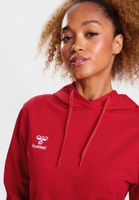 Rote Kapuzenjacke mit einer vorderen Tasche, die ein weißes gesticktes Logo auf der Brust aufweist. Der Stoff wirkt glatt, mit einem klassischen Design.