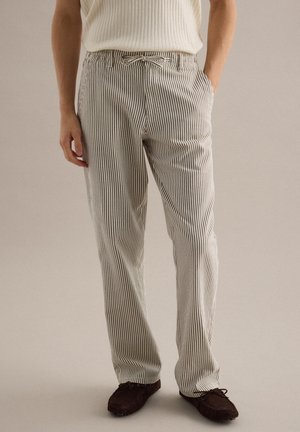 Person, die weiße und schwarze vertikal gestreifte Hosen mit einem Taillengürtel und dunkelbraunen Schnürschuhen trägt, steht mit einer Hand in der Tasche.