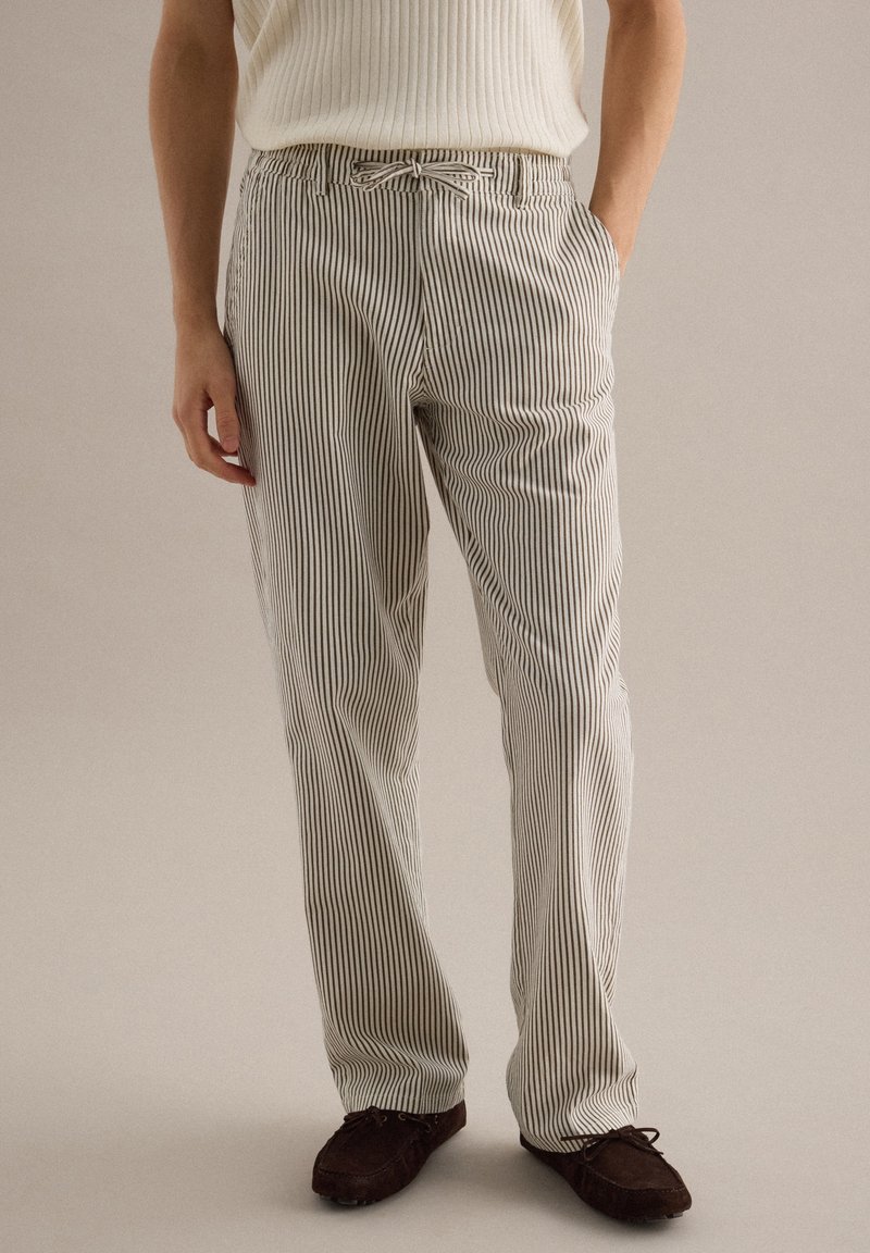 Persona vistiendo pantalones a rayas verticales blancas y negras con cinturón de lazo y zapatos marrón oscuro con cordones, de pie con una mano en el bolsillo.