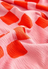 Puha, texturált felületű textil, nagy, szabálytalan vörös négyzetekkel, világos rózsaszín háttéren, amely élénk színkontrasztot teremt.