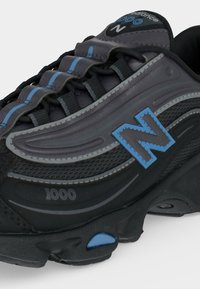 Μαύρο και γκρι αθλητικό παπούτσι New Balance 1000 με μπλε λεπτομέρειες και πάνελ από δίχτυ, εμφανίζεται σε κοντινή πλευρική λήψη.