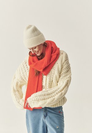 Dikke gebreide kabeltruicardigan, levendig rode geribde sjaal en lichtgrijze beanie. Zachte texturen, ontspannen pasvorm, met een knoopdetail op de cardigan.