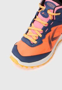 Buty sportowe z siateczkową cholewką w jaskrawym pomarańczowym i różowym kolorze, z granatowymi akcentami, żółtymi sznurowadłami oraz teksturowaną gumową podeszwą zapewniającą przyczepność.