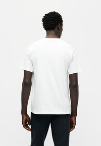 Λευκό κοντομάνικο μπλουζάκι από βαμβακερό ύφασμα, με στρογγυλή λαιμόκοψη και ίσια τελείωμα, φαίνεται από πίσω. Απλός, χωρίς διακοσμήσεις σχεδιασμός.