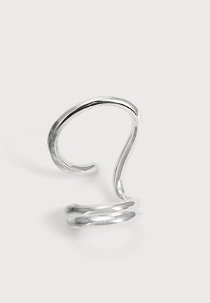 Une bague ouverte en argent poli, de type manchette, avec un anneau incurvé aux formes sculpturales et deux boucles à la base, présentée sur un fond clair.