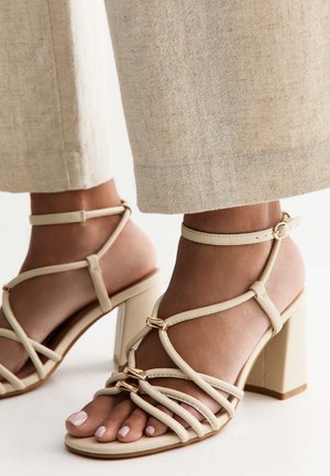 Voeten met beige sandalen met blokhak en bandjes gecombineerd met lichtbeige wijde broekspijpen tegen een witte achtergrond.