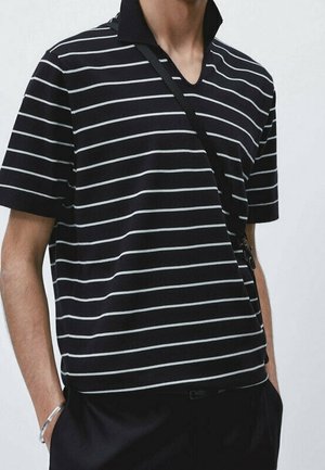 Person wearing black short-sleeve polo with white horizontal stripes and a black crossbody bag strap, hands in black pants pockets.