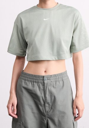 Osoba ubrana w jasną zieloną krótką siateczkową koszulkę marki Nike i luźne szare spodnie, stojąca z rękami swobodnie opuszczonymi wzdłuż ciała na jednolitym tle.