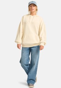 Crème hoodie met reliëf "BILLABONG" logo, kangoeroezak, oversized pasvorm, gecombineerd met lichtblauwe wijde jeans en een denim pet.