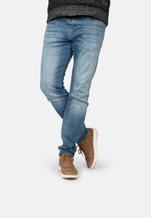 Jeans de mezclilla azul claro con un ajuste Slim, caracterizados por áreas desgastadas y un diseño clásico de cinco bolsillos, combinados con zapatillas marrones.
