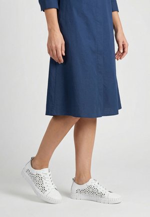 Frau, die in einem blauen knielangen Kleid weiße Sneakers mit Spitzenmuster und Schnürsenkeln vor einem einfarbigen Hintergrund trägt.