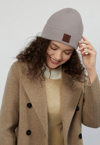 Harmaa ribbikuvioinen neulepipo, jossa on ruskea nahkalaatta; yhdistettynä vaaleanruskeaan kaksiriviseen takkiin ja vaaleanbeigeen neuleeseen.