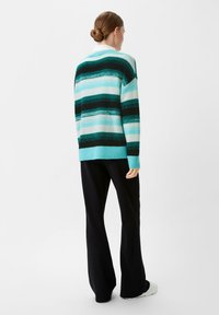 Knitted sweater with teal, blue, and black horizontal stripes, featuring a relaxed fit and ribbed cuffs. Paired with black pants.