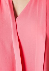 Close-up af en person iført et lyst pink struktureret tøjstykke med overlappende stofpaneler nær halsudskæringen.