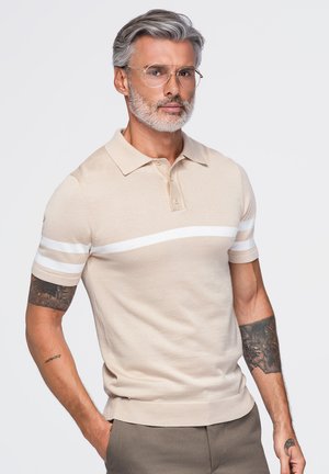Homme aux cheveux gris et à la barbe portant des lunettes, un polo beige à rayures blanches et un pantalon marron, debout les mains dans les poches.