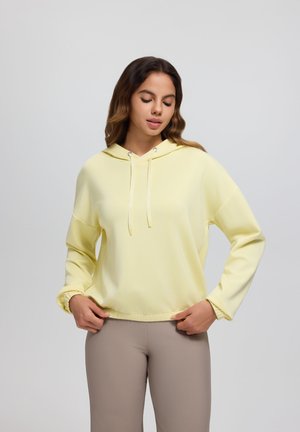 Junge Frau mit geschlossenen Augen, die einen hellgelben Hoodie und beige enganliegende Hosen trägt und vor einem einfarbigen, hellgrauen Hintergrund steht.