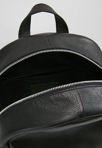 Svart texturerat läderryggsäck med öppen silverdragkedja som visar en tom huvudfack och ett handtag på toppen.