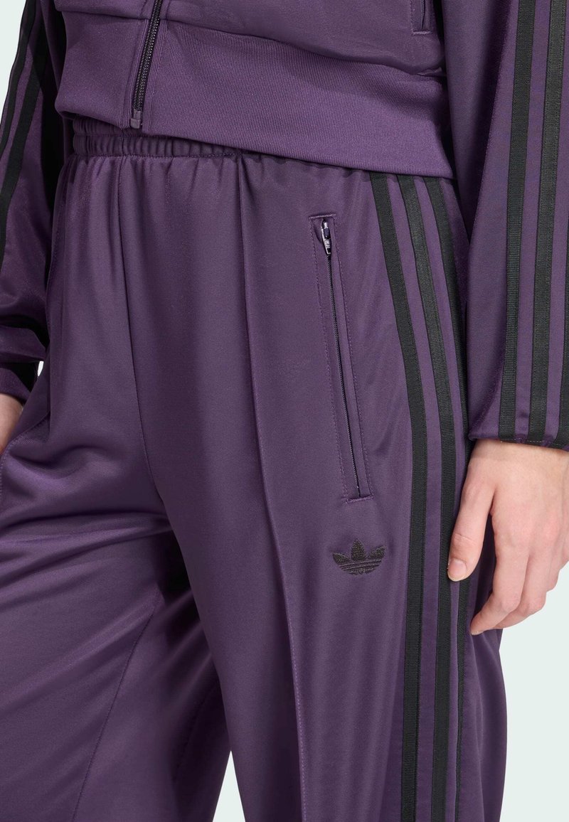 Survêtement de sport violet avec des bandes noires Adidas, poche zippée et logo embossé sur le bas de la jambe, en matériau lisse et élégant.