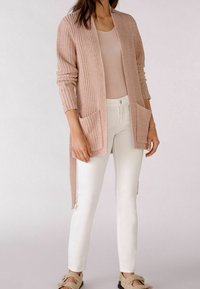 Cárdigan de punto rosa claro con abertura frontal, patrón texturizado, bolsillos laterales y mangas largas, combinado con pantalones blancos y zapatillas beige.