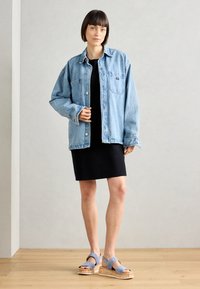Giacca in denim di colore azzurro chiaro con due tasche frontali, abbinata a un vestito nero e sandali con piattaforma blu e suole in sughero.