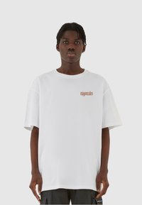 Hvid oversized T-shirt lavet af bomuld, med rund halsudskæring og et lille trykt logo i brun på venstre side. Simpelt design.