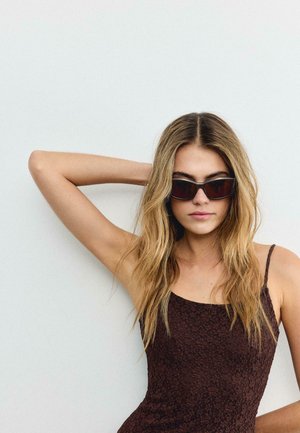 Mladá žena s dlouhými blond vlasy, oblečená v tmavých květovaných šatech a slunečních brýlích, pózující s jednou rukou ohnutou za hlavou na jednobarevném pozadí.