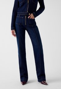 Donkere blauwe denim jumpsuit met een rits aan de voorkant, zakken en gouden accenten. Heeft rechte pijpen en contrasterende stiksels voor extra detail.