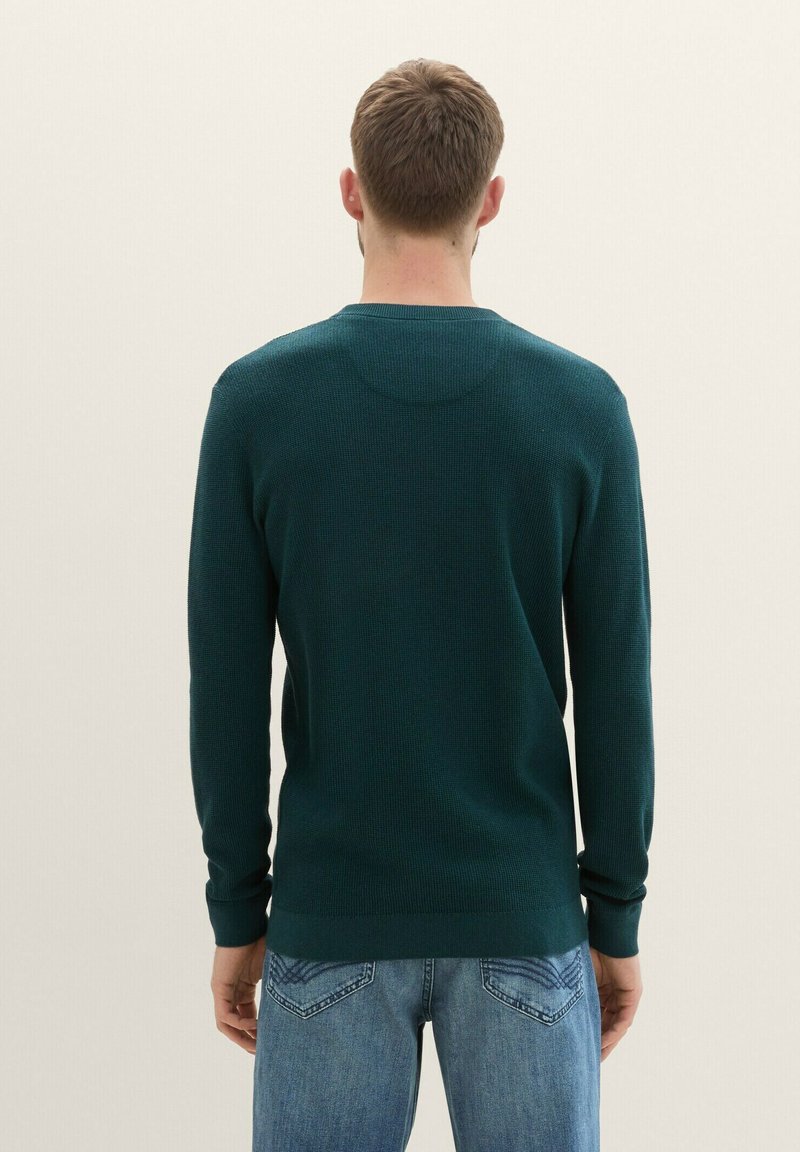 Dunkelgrüner, gerippter Strickpullover mit langen Ärmeln und rundem Halsausschnitt, der einen taillierten Saum und kontrastierende Texturen aufweist.
