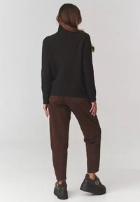 Kobieta stoi, ubrana w czarny prążkowany sweter z golfem, brązowe spodnie i czarne trampki na platformie, zwrócona plecami do kamery.