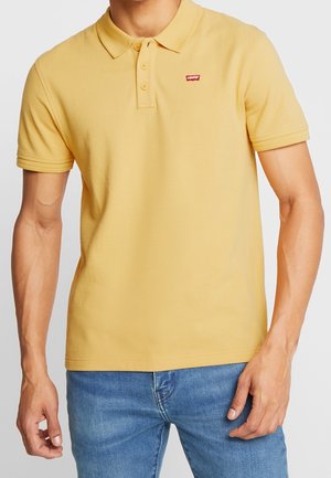 Mężczyzna ubrany w żółtą koszulkę polo z krótkim rękawem, z trzema guzikami i czerwonym logo Levi's, w niebieskich dżinsach, stojący na jednolitym tle.