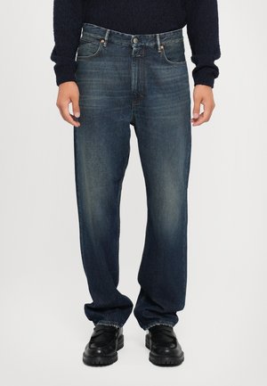 Personne portant un jean bleu foncé délavé à jambes droites, des chaussures noires et un pull foncé à manches longues, debout devant un fond uni.