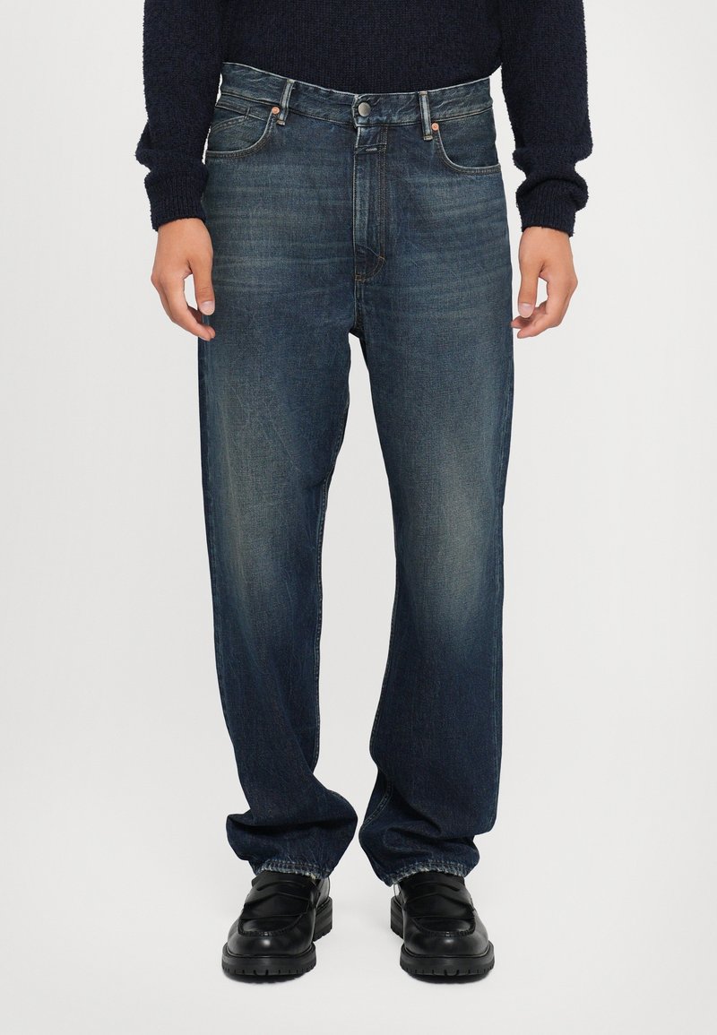 Personne portant un jean bleu foncé délavé à jambes droites, des chaussures noires et un pull foncé à manches longues, debout devant un fond uni.