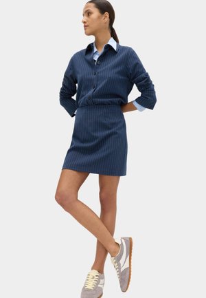 Kobieta stojąca z rękami za plecami, ubrana w granatową spódnicę w prążki i dopasowany, zapinany na guziki kardigan na jasnoniebieskiej koszuli, w sportowych butach.