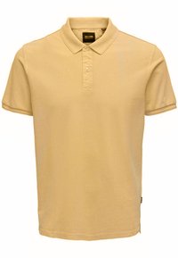 Camisa polo beige con cuello clásico, tapeta de tres botones, mangas cortas y material de algodón suave, con una pequeña etiqueta de logo en el costado.