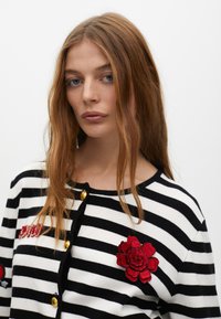 Czarny i biały sweter w paski z okrągłym dekoltem, złotymi guzikami i czerwonym haftowanym kwiatem po lewej stronie klatki piersiowej. Miękka tekstura dzianiny.