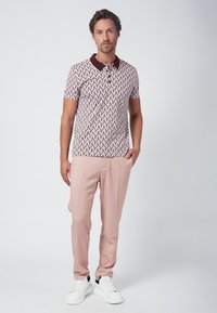 Kortärmad polo med ett geometriskt mönster i vitt, vinrött och blått, i kombination med ljusrosa byxor och vita sneakers.
