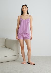 Randig rosa och vit sovdräkt med en v-ringad topp och shorts. Modell som bär fluffiga tofflor, står på en neutral matta.