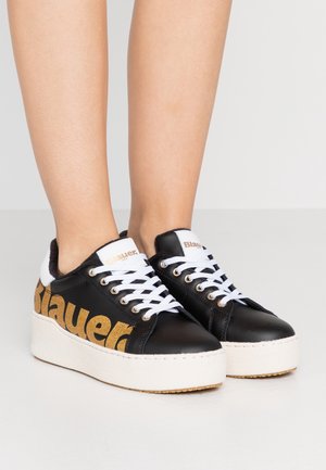 Baskets plateformes noires avec lacets blancs, marquage doré « Blauer » sur le côté, portées sur une paire de jambes sur un fond uni.