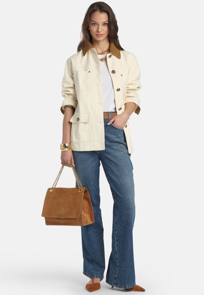 Hellbeige Jacke mit braunem Kragen, weißem Oberteil, blauen ausgestellten Jeans und brauner Wildleder-Handtasche. Accessoires umfassen eine goldene Uhr und ein Armband.