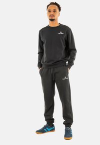 Sudadera de algodón negra y pantalones jogger con puños elásticos, con un logo en el pecho y en los pantalones. Estilizados con zapatillas de deporte con acentos azules.