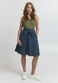 Jupe en denim avec une taille élastique et un détail de nœud, associée à un débardeur vert et des baskets blanches. Texture lisse, design arrivant au genou.