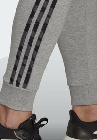 Grå sweatpants med tre svarta randiga detaljer längs sidan, ribbade muddar och en slät textur. Kombineras med en svart sneaker.