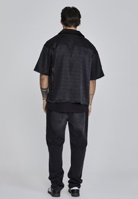 Camisa negra de manga corta con un patrón texturizado, combinada con jeans negros y zapatos oscuros, vista desde atrás contra un fondo neutro.